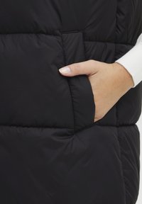 Schwarze Steppweste mit einem gesteppten Design, Seitentasche und einer glatten, glänzenden Textur. Der Stoff wirkt leicht und wasserabweisend.