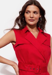 Czerwona sukienka bez rękawów z kołnierzykiem, z paskiem w talii i guzikami. Gładki materiał z dopasowanym krojem.