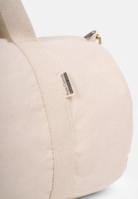 Round canvas bag in cream color with a textured fabric, featuring strong woven handles and a plastic-free label attached.