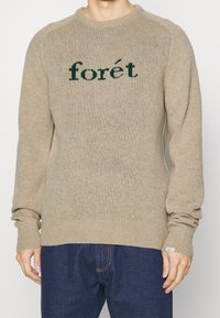 Beżowy sweter dzianinowy z zielonym haftem "forét" na piersi, ściągacze przy mankietach i dolnym brzegu, okrągłym dekoltem oraz fakturą materiału.