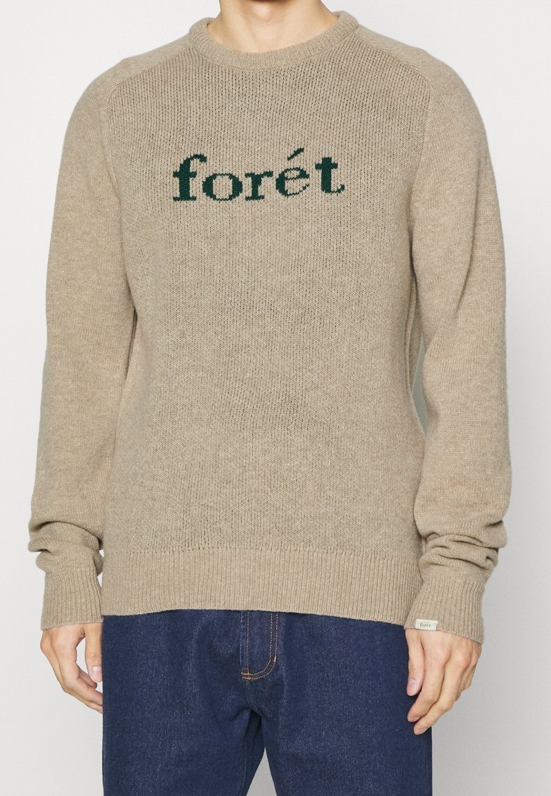 Beżowy sweter dzianinowy z zielonym haftem "forét" na piersi, ściągacze przy mankietach i dolnym brzegu, okrągłym dekoltem oraz fakturą materiału.