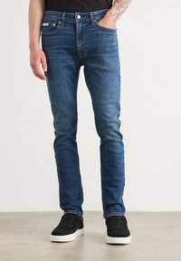 Mees, kes kannab slim-fit siniseid teksaseid mustade tossudega, üks käsi taskus ja nähtav tätoveering alakaenal, lihtsa valge tausta ees.