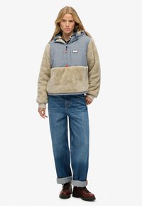 Kurtka typu pullover z szarym nylonowym górnym częścią, beżowym dolnym z polaru i kapturem ze sznurkiem. Połączona z podwiniętymi niebieskimi dżinsami i brązowymi butami.