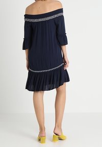 Robe bleu marine à épaule nue avec bordure brodée, manches trois-quarts, ourlet volanté et texture fluide, associée à des chaussures jaunes à talons blocs.