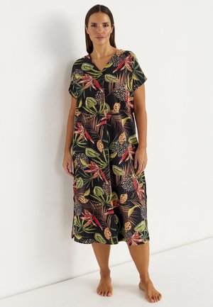 Kobieta stoi boso w czarnej sukience do kolan z czerwonymi, zielonymi i beżowymi tropikalnymi wzorami liści i kwiatów.
