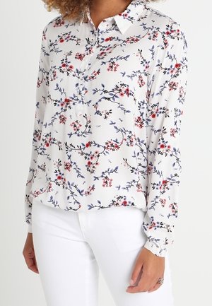 Weiße Bluse mit langen Ärmeln und buntem Blumenmuster, vorne geknöpft und mit Kragen, getragen zu weißen Hosen.