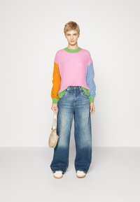 Pull surdimensionné coloré avec des manches côtelées roses, orange, bleues et vertes ; jean large en denim clair ; chaussures blanches ; sac à main beige.