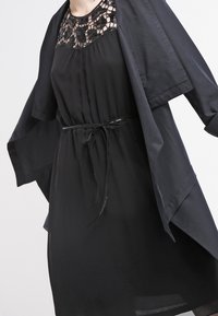 Robe noire avec un col en dentelle et un superposé fluide à manches. Dispose d'une ceinture à la taille pour un ajustement réglable, fabriquée en tissu léger.