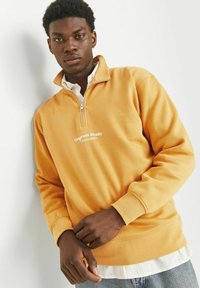 Felpa gialla con zip a quarto, con polsini e orlo a costine, caratterizzata da un piccolo logo bianco sul petto, indossata sopra una camicia bianca con colletto.