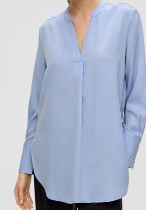 Femme portant un chemisier bleu clair à manches longues avec une fente en V et des poignets boutonnés, associé à un pantalon noir.