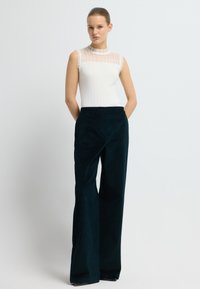 Bílý top bez rukávů s žebrovanou texturou a průhlednými vertikálními detaily. Tmavě zelené široké kalhoty, hladká látka a viditelné boční kapsy.