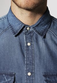 Farkkupaita, jossa on terävä kaulus, kaksi rintataskua ja hopeiset napit; keskisinisessä sävyssä, teksturoidulla kangaspinnalla.