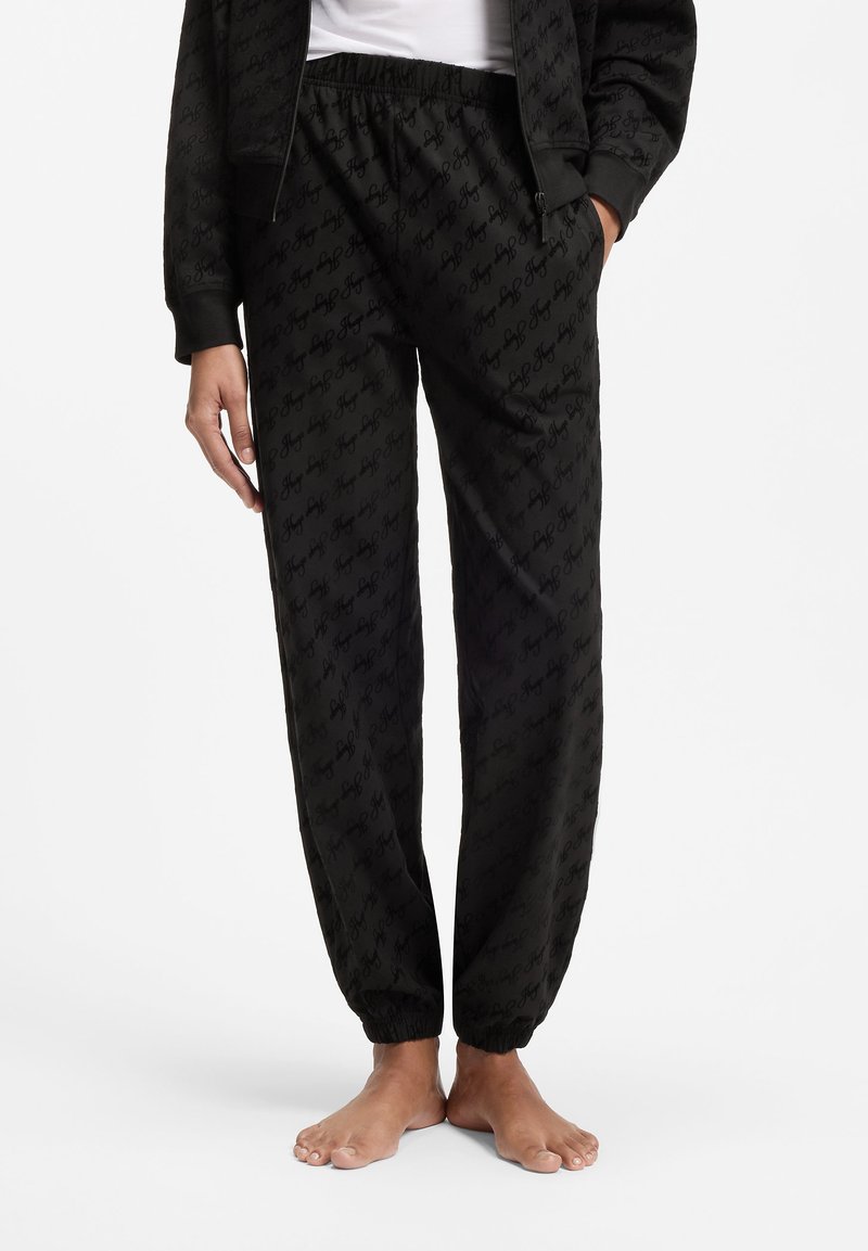 Personne portant un pantalon noir à taille élastique avec un motif subtil en écriture répétée, des poignets élastiques, les mains dans les poches, pieds nus sur un sol blanc.