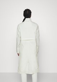 Osoba s pletenými vlasy, oblečená v dlouhém bílém trench coat, viděná zezadu na jednoduchém světlém pozadí.