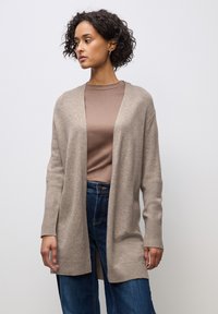 Béžový svetr vyrobený z měkké tkaniny, s otevřeným předním dílem, dlouhými rukávy a žebrovanými manžetami. Nosený přes hnědý top a modré džíny.