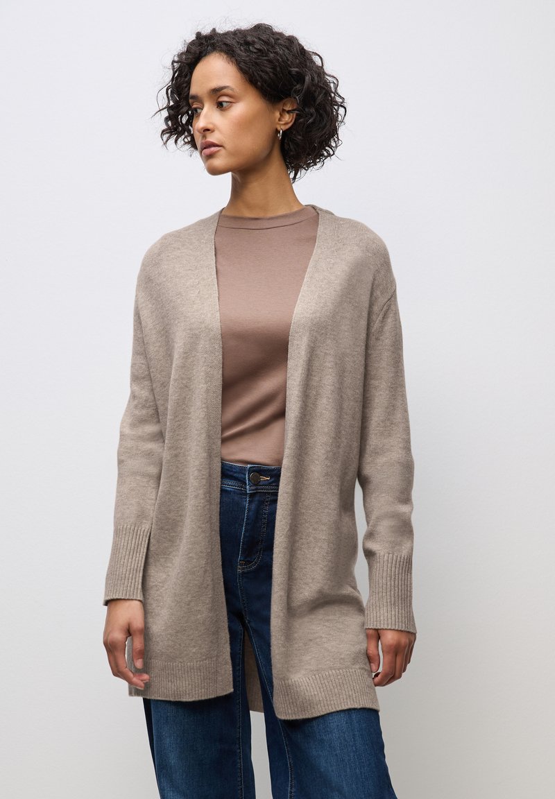 Béžový svetr vyrobený z měkké tkaniny, s otevřeným předním dílem, dlouhými rukávy a žebrovanými manžetami. Nosený přes hnědý top a modré džíny.