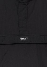 Sort stof med en glat tekstur, der har et synligt logo-mærke. Enkelt design og lige snit med syninger langs kanterne.