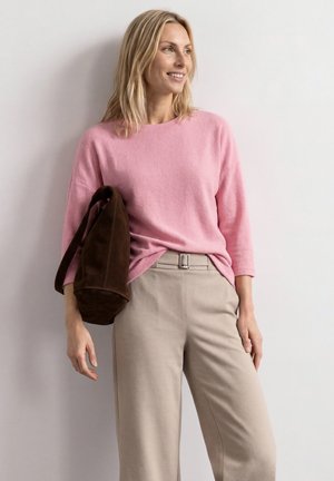 Femme en pull rose et pantalon beige tenant un sac marron foncé sur l'épaule, debout et souriante devant un mur blanc.
