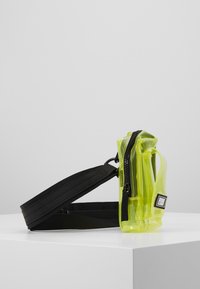 Transparent gul crossbody-veske med et svart justerbart stroppe, med glidelås og en rektangulær logo-patch. Glatt tekstur.