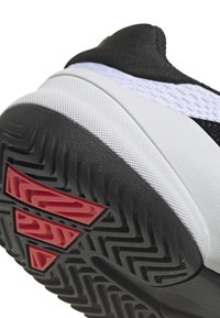 Black athletic shoe featuring a textured surface, white midsole, and red accents on the sole. Tread pattern is designed for traction.