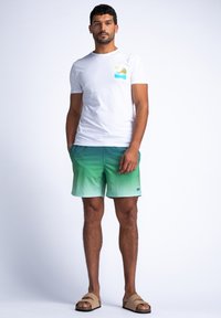 Hvit t-skjorte med et fargerikt stranddesign, kombinert med grønne gradiente badebukser og brune sandaler, stående på en ensfarget bakgrunn.
