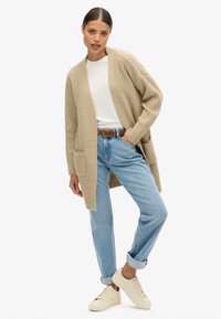 Beige strikket cardigan med åben front og lommer, båret over en hvid t-shirt og lyseblå opkrøllede jeans. Hvide sneakers.