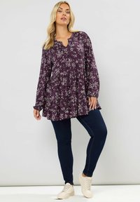 Violetti kukallinen pitkähiha Tunika, jossa on frillahelma, yhdistettynä tummanssinisiin slim-fit housuihin ja vaaleisiin nauhakenkiin.