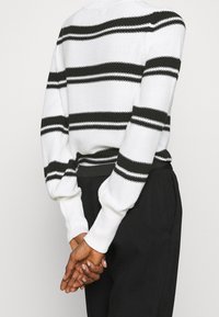 Strickpullover mit weißem Körper und schwarzen horizontalen Streifen. Gerippte Bündchen und Saum sorgen für Textur und werden mit schwarzen Hosen kombiniert.