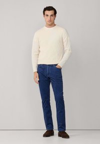 Suéter de punto trenzado color crema, pantalones azules, zapatos marrones. Diseño simple con un patrón texturizado en el suéter y un corte recto en los pantalones.