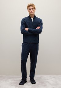 Chaqueta de punto azul marino con pantalones a juego, hechos de una tela suave y ligera. El modelo está de pie con los brazos cruzados, usando zapatillas deportivas oscuras.