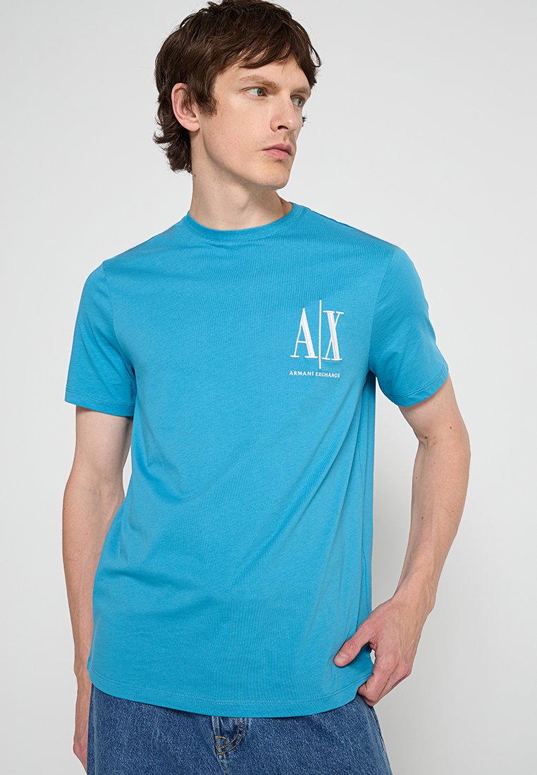 Młody mężczyzna ubrany w jasnoniebieską koszulkę Armani Exchange z białym logo na piersi, stojący na tle gładkiego białego tła.