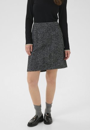 Femme portant un haut noir à manches longues, une jupe grise à motif pied-de-poule, des chaussettes grises et des mocassins noirs brillants, se tenant devant un fond neutre.