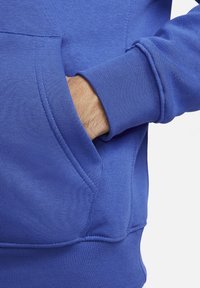 Felpa in cotone blu con tasca a marsupio, polsini a coste e una consistenza morbida, con la mano parzialmente all'interno della tasca.