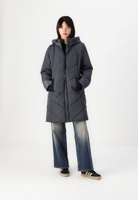 Manteau matelassé gris foncé avec une capuche, doté d'une fermeture éclair à l'avant, d'un motif en chevrons et de poches latérales, associé à un jean évasé et à des baskets.