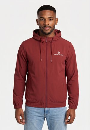 Homme portant une veste à capuche zippée bordeaux avec le logo Sergio Tacchini, associée à un jean bleu clair, debout devant un fond uni.