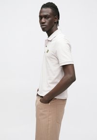 Lichtroze poloshirt met een geel logo, gecombineerd met beige geribbelde broek. Voorzien van een klassieke kraag en korte mouwen.