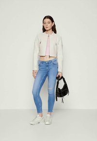 Casaco branco cropped com detalhes de fecho prata, combinado com jeans skinny azul claro com um joelho rasgado. O modelo segura uma bolsa preta.