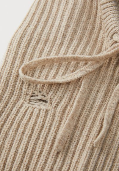 Tissu tricoté beige avec des côtes verticales, présentant un détail à nouer et une petite découpe horizontale. Texture douce et couleur naturelle.