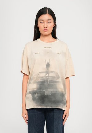 T-shirt oversize beige avec un graphique dégradé d'une femme sur une voiture, comportant les textes "DON DUP" et "FABRIQUÉ EN ITALIE". Tissu en coton doux.