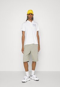 Hvid polo t-shirt med korte ærmer, gul cap, beige shorts og hvide sneakers. Har logo-broderi på skjorten og flere teksturer.