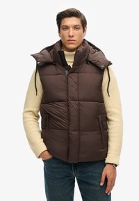 Braune Puffer-Weste mit hohem Kragen und Kapuze, ausgestattet mit einer gesteppten Textur, einem Frontreißverschluss und seitlichen Taschen. Getragen über einem cremfarbenen Strickpullover.