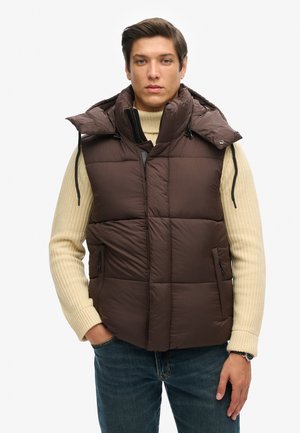 Braune Puffer-Weste mit hohem Kragen und Kapuze, ausgestattet mit einer gesteppten Textur, einem Frontreißverschluss und seitlichen Taschen. Getragen über einem cremfarbenen Strickpullover.