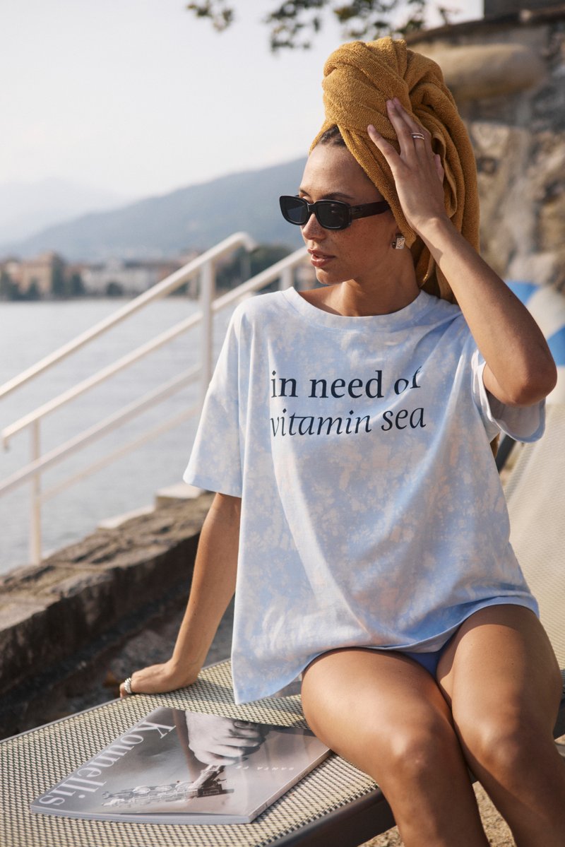 T-shirt surdimensionné bleu tie-dye avec le texte "besoin de vitamine mer". Le mannequin porte des lunettes de soleil et une serviette jaune sur la tête, assise près de l'eau.