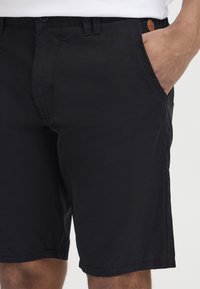 Shorts en coton noirs avec une texture lisse, dotés de poches latérales, d'une fermeture à bouton et d'un petit logo sur la ceinture.