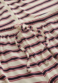 Tkanina krem boje s prugama u mornarsko plavoj i crvenoj boji, s vrpcama za vezanje i gumbima duž ovratnika za podešavanje.