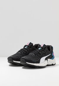 Une paire de chaussures de course noires Puma avec des semelles texturées blanches, une bande latérale blanche et des accents roses et bleus, présentée sur une surface blanche contre un arrière-plan gris.