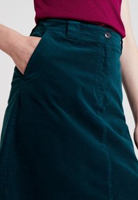 Jupe teal avec une poche latérale, texture lisse, fermeture à boutons et un design ajusté. Présente une couture visible le long du côté.