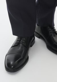 Chaussures de ville en cuir noir avec une finition lisse, bout pointu et lacets. Portées avec un pantalon texturé sombre, mettant en valeur un style formel.