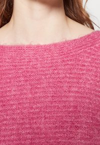Pull rose duveteux avec un motif tricoté texturé, un large décolleté et une apparence douce. Vue en gros plan mettant en valeur la qualité du matériau.