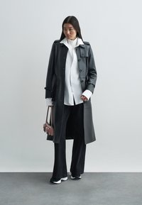 Schwarzer Ledermantel mit Knöpfen, über einem weißen Hemd getragen. Kombiniert mit weiten schwarzen Hosen und Sneakers mit weißen Akzenten. Kleine rosa Tasche.